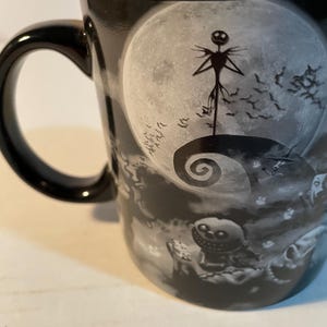May include: A black ceramic mug featuring a monochromatic graphic print of a tall, thin character standing on a spiral hill against a large full moon, with a small round character holding dice in the foreground and a flock of bats in the sky.