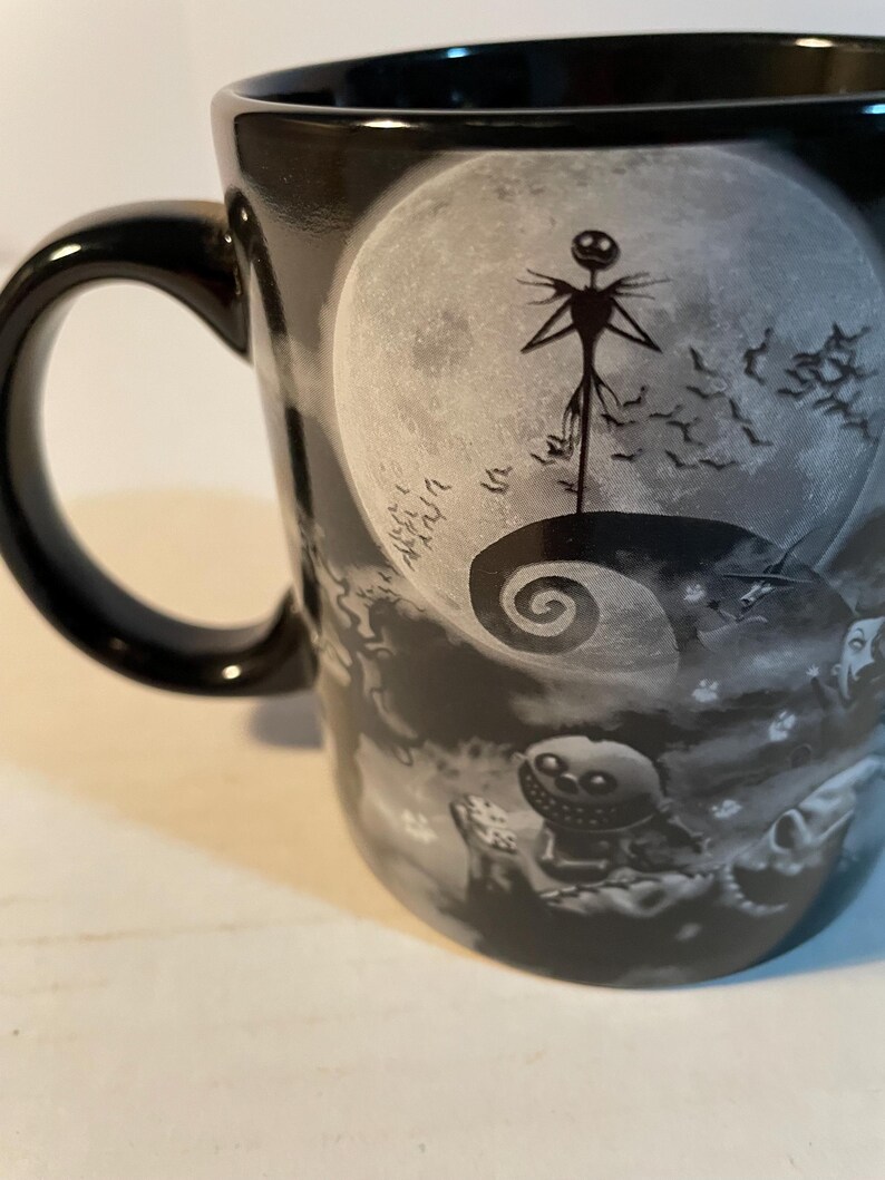 May include: A black ceramic mug featuring a monochromatic graphic print of a tall, thin character standing on a spiral hill against a large full moon, with a small round character holding dice in the foreground and a flock of bats in the sky.