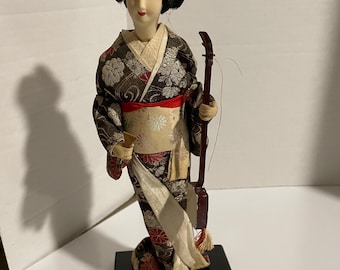 Pode incluir: Uma boneca gueixa japonesa, adornada com um quimono tradicional com padrões florais em vermelho, dourado e castanho. A boneca tem um penteado preto e segura um instrumento de cordas. Está sobre uma base retangular preta.