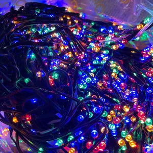 May include: A dense cluster of tangled string lights featuring small, multi-colored LED bulbs in red, blue, green, and yellow. The lights are connected by a black wire and are partially contained within a translucent, crinkled plastic bag.