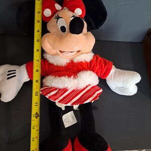May include: A red and white plush Minnie Mouse wearing a red and white candy cane striped skirt and a red coat with white fur trim. She is wearing a red bow with white polka dots on her head.
