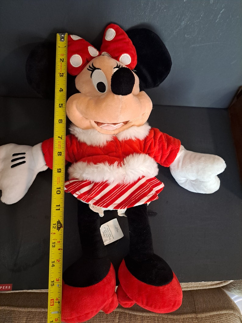 May include: A red and white plush Minnie Mouse wearing a red and white candy cane striped skirt and a red coat with white fur trim. She is wearing a red bow with white polka dots on her head.