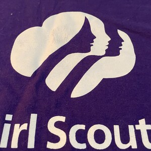 May include: A purple t-shirt featuring a white graphic of three overlapping female silhouettes. Below the graphic, the text reads "Girl Scout" and "ncil of the Nation's Ca". The design is screen-printed in white ink on the front of the purple fabric.