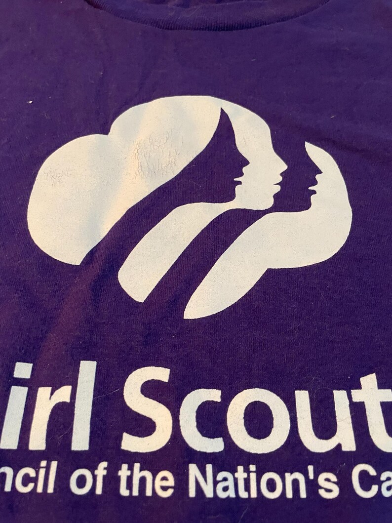 May include: A purple t-shirt featuring a white graphic of three overlapping female silhouettes. Below the graphic, the text reads "Girl Scout" and "ncil of the Nation's Ca". The design is screen-printed in white ink on the front of the purple fabric.