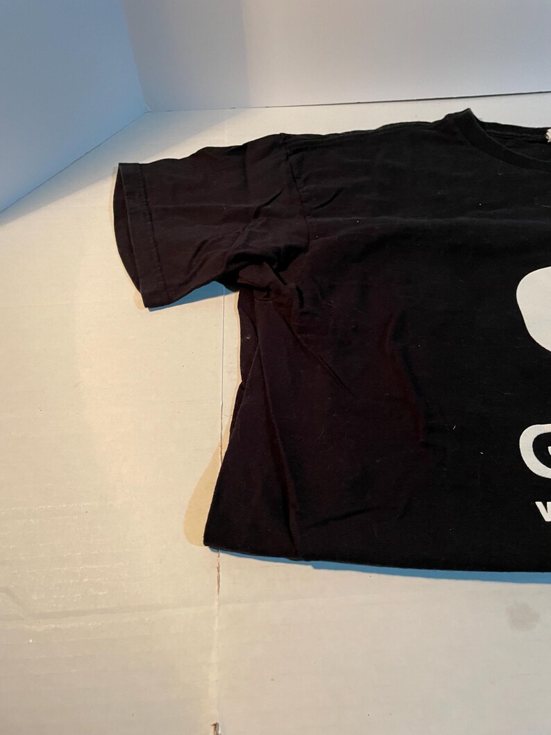 May include: A black short-sleeved t-shirt featuring a large white graphic print on the front. The design includes a partial white shape at the top and the white capital letters G and V visible near the bottom edge of the fabric.