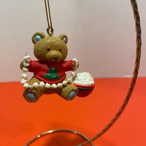 May include: A brown teddy bear Christmas ornament wearing a red sweater with a green Christmas tree design. The bear is holding a white garland and a red and white ornament. The ornament is hanging from a gold-colored wire.
