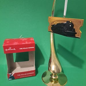 May include: A Hallmark Christmas tree ornament featuring a black train with a gold ticket-shaped frame. The ornament is in its original packaging, which is red with white snowflakes and the words "The Polar Express".