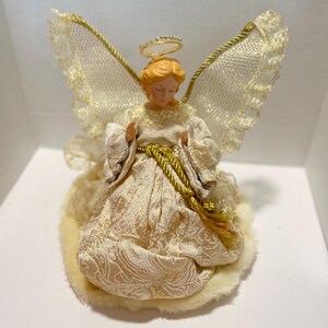 May include: A decorative angel figurine with a halo and wings. The angel is dressed in a cream-colored gown with gold accents and a gold rope belt. The wings are made of lace and gold trim. The figurine is sitting on a fluffy, cream-colored base.