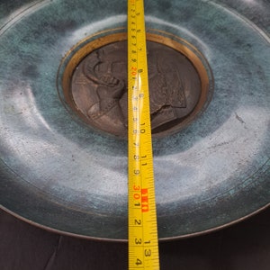 May include: A round, antique bronze plate with a raised relief of a profile portrait. The plate has a verdigris finish and a gold-colored inner ring. The plate's diameter is approximately 11 inches.