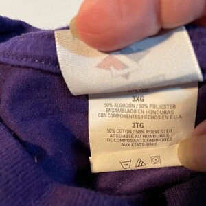 May include: A purple garment with a white interior clothing tag. The tag displays the size 3XG and 3TG, and states 50% Algod&oacute;n/50% Poliester, 50% Coton/50% Polyester, made in Honduras with components from the U.S.A., including laundry care symbols.
