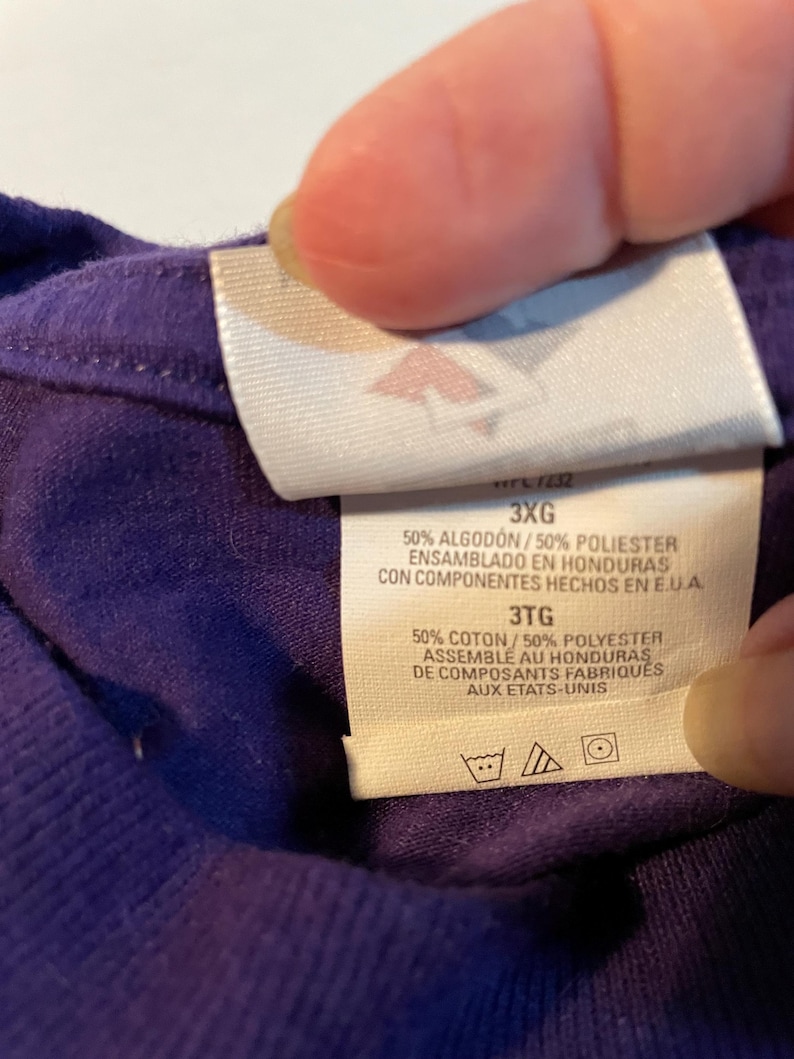 May include: A purple garment with a white interior clothing tag. The tag displays the size 3XG and 3TG, and states 50% Algod&oacute;n/50% Poliester, 50% Coton/50% Polyester, made in Honduras with components from the U.S.A., including laundry care symbols.