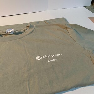 May include: A sage green crew neck t-shirt featuring the white printed text Girl Scouts Leader and a small trefoil logo on the upper left chest area. The shirt has a standard short sleeve design and a ribbed collar with a white interior neck tag.