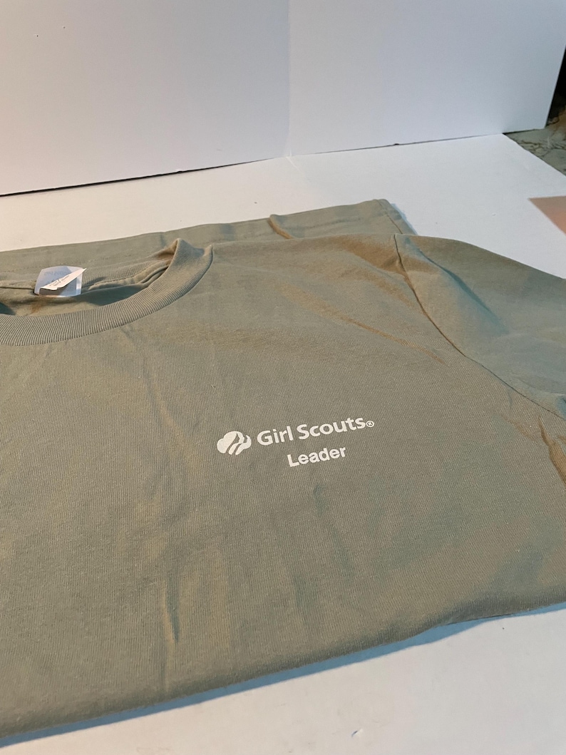 May include: A sage green crew neck t-shirt featuring the white printed text Girl Scouts Leader and a small trefoil logo on the upper left chest area. The shirt has a standard short sleeve design and a ribbed collar with a white interior neck tag.