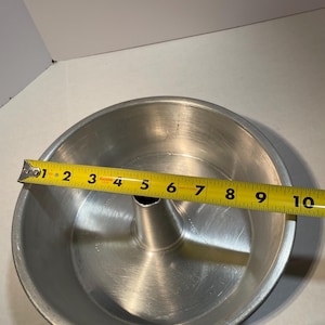 May include: A silver-coloured, round angel food cake tin with a central tube. The tin is approximately 25 cm in diameter, as measured by a yellow tape measure. The tin is made of metal and has a shiny finish.