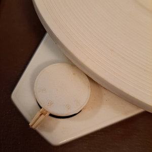 Pu&ograve; includere: Primo piano di un giradischi bianco con una base in legno marrone. Il giradischi ha un piatto bianco e una manopola di controllo della velocit&agrave; con i numeri 33, 45 e 78.