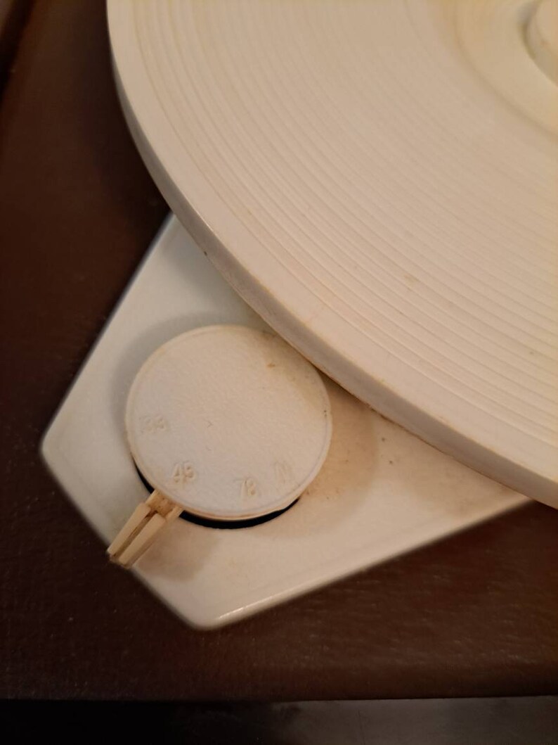 Pu&ograve; includere: Primo piano di un giradischi bianco con una base in legno marrone. Il giradischi ha un piatto bianco e una manopola di controllo della velocit&agrave; con i numeri 33, 45 e 78.