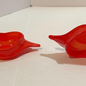 May include: Two red, heart-shaped glass bowls with wing-like extensions. The bowls are glossy and appear to be made of a translucent material. The bowls are displayed on a white surface.