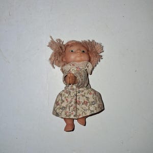 May include: A vintage doll with a peach-colored face, green eyes, and pink yarn hair styled in two side buns. The doll wears a floral print dress with a lace collar. The doll's arms are raised, and its feet are visible.
