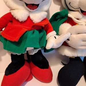 Puede incluir: Dos personajes de peluche de Mickey Mouse con tem&aacute;tica navide&ntilde;a. Uno est&aacute; vestido con un vestido rojo y verde con un cuello blanco y zapatos rojos. El otro lleva un traje de Pap&aacute; Noel rojo y blanco con una bolsa verde y zapatos negros.