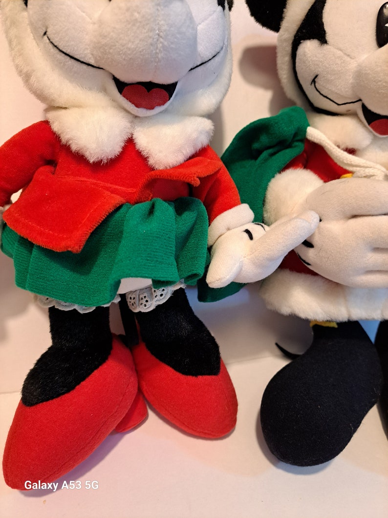 Puede incluir: Dos personajes de peluche de Mickey Mouse con tem&aacute;tica navide&ntilde;a. Uno est&aacute; vestido con un vestido rojo y verde con un cuello blanco y zapatos rojos. El otro lleva un traje de Pap&aacute; Noel rojo y blanco con una bolsa verde y zapatos negros.