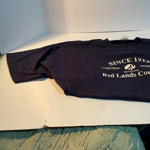 K&ouml;nnte beinhalten: Ein dunkelblaues T-Shirt mit kurzen &Auml;rmeln und wei&szlig;em Aufdruck, der SINCE 1912, ein Girl Scouts-Logo und die Worte Red Lands Cou zeigt. Das T-Shirt hat einen Rundhalsausschnitt und besteht aus einfarbigem Stoff mit einer Standardpassform.