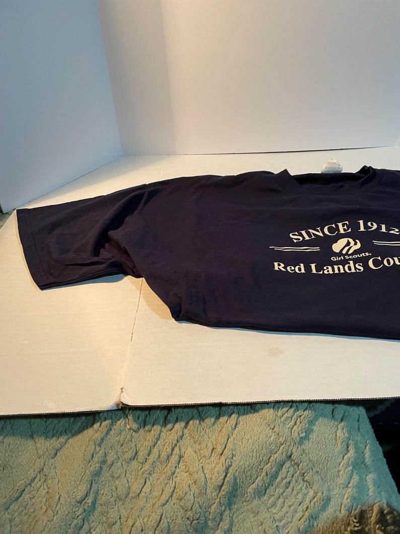 K&ouml;nnte beinhalten: Ein dunkelblaues T-Shirt mit kurzen &Auml;rmeln und wei&szlig;em Aufdruck, der SINCE 1912, ein Girl Scouts-Logo und die Worte Red Lands Cou zeigt. Das T-Shirt hat einen Rundhalsausschnitt und besteht aus einfarbigem Stoff mit einer Standardpassform.