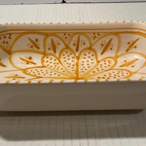 May include: A rectangular ceramic serving dish with an octagonal rim, featuring a white base color and a hand-painted yellow floral pattern on the interior. The design includes intricate yellow dots and lines arranged in a symmetrical petal motif.