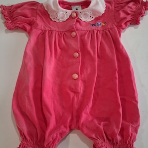 May include: A bright pink romper featuring short puffed sleeves with elastic cuffs, a white scalloped collar with eyelet embroidery, and four rose-shaped buttons down the front. A small floral embroidery detail is visible on the left chest.