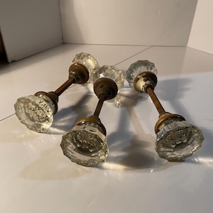 May include: Four antique glass doorknobs with faceted crystal-like designs. The knobs have aged brass-colored metal stems and bases, showing signs of wear. The clear glass reflects light, creating a sparkling effect. These vintage knobs are ideal for restoration projects.