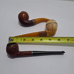 Op de afbeelding: Twee vintage houten pijpen. E&eacute;n pijp heeft een bruine kom en een gele steel. De andere pijp heeft een bruine kom en een zwarte steel. Een liniaal is op de achtergrond te zien.