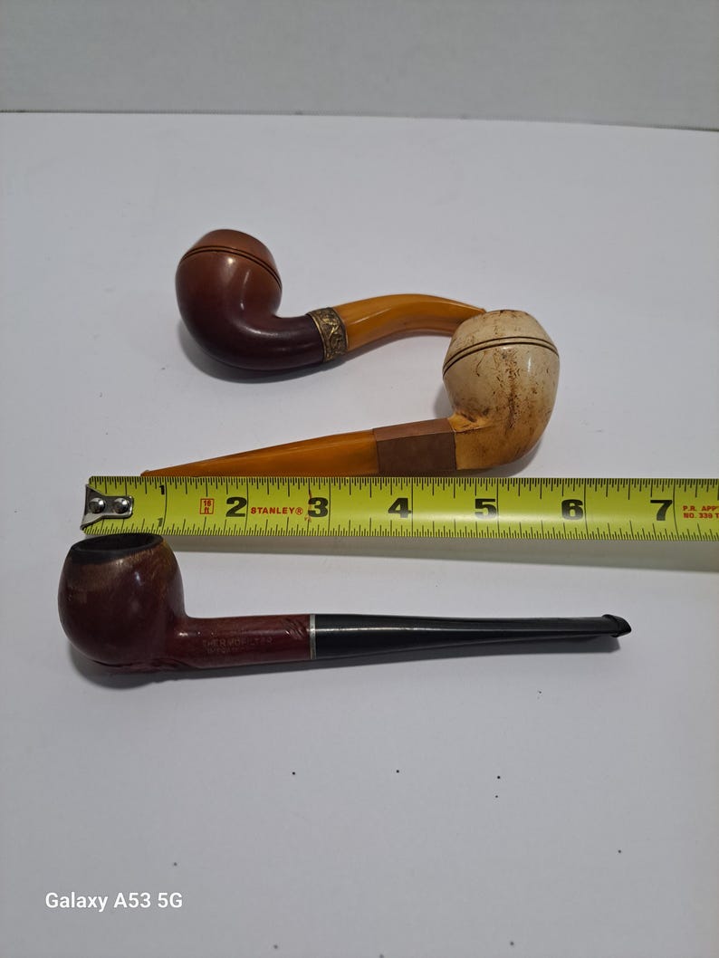 Op de afbeelding: Twee vintage houten pijpen. E&eacute;n pijp heeft een bruine kom en een gele steel. De andere pijp heeft een bruine kom en een zwarte steel. Een liniaal is op de achtergrond te zien.