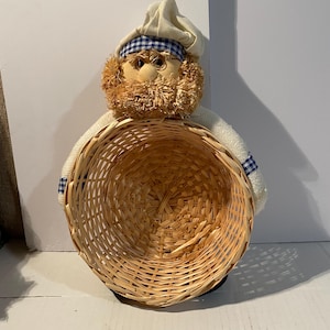May include: A decorative chef figurine featuring a tan fabric face with a straw beard, wearing a white chef hat with a blue and white checkered band. The figure has white sleeves with blue and white checkered cuffs and holds a round woven wicker basket.