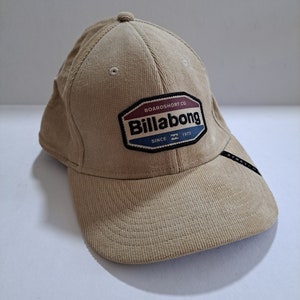 May include: A beige corduroy baseball cap with a black and red Billabong logo patch. The patch reads "Boardshort Co. Billabong Since 1973".