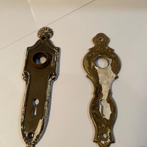 May include: Two antique door hardware pieces. One is a rectangular escutcheon with a keyhole and a decorative top. The other is an oval-shaped escutcheon with a keyhole. Both have a distressed, aged appearance.