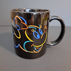 Peut inclure: Mug en c&eacute;ramique noire avec un motif de personnage de dessin anim&eacute; color&eacute;. Le motif comprend des contours bleus, orange et jaunes d'un visage et d'un n&oelig;ud. La tasse a une finition brillante et une anse incurv&eacute;e.