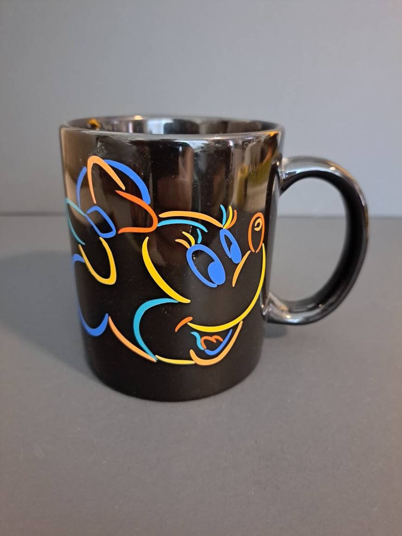 Peut inclure: Mug en c&eacute;ramique noire avec un motif de personnage de dessin anim&eacute; color&eacute;. Le motif comprend des contours bleus, orange et jaunes d'un visage et d'un n&oelig;ud. La tasse a une finition brillante et une anse incurv&eacute;e.
