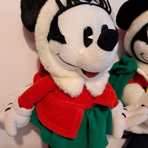 Puede incluir: Un juguete de peluche de Minnie Mouse con un abrigo rojo y una falda verde. Tiene ojos negros y pesta&ntilde;as. El juguete est&aacute; de pie sobre una superficie blanca.