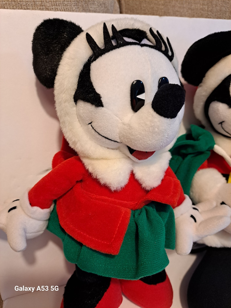 Puede incluir: Un juguete de peluche de Minnie Mouse con un abrigo rojo y una falda verde. Tiene ojos negros y pesta&ntilde;as. El juguete est&aacute; de pie sobre una superficie blanca.