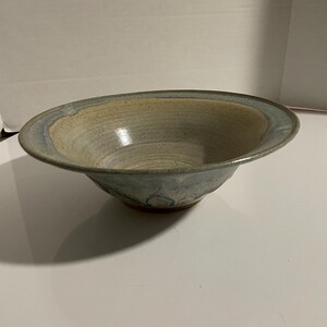 May include: A handmade ceramic bowl with a wide, shallow design. The bowl features a light beige interior and a blue-grey rim. The exterior has a natural, earthy tone. This decorative bowl is suitable for serving or display.