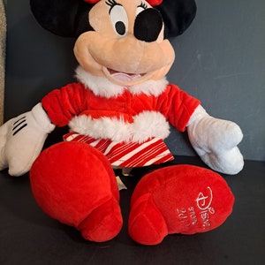 May include: Red and white plush Minnie Mouse wearing a red and white striped skirt and a red coat with white fur trim. She has a red bow with white polka dots on her head. The plush toy has the text "Disney Store" and "2017" on the bottom of one foot.