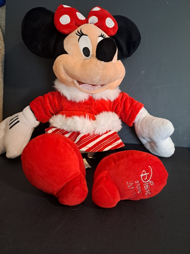 May include: Red and white plush Minnie Mouse wearing a red and white striped skirt and a red coat with white fur trim. She has a red bow with white polka dots on her head. The plush toy has the text "Disney Store" and "2017" on the bottom of one foot.