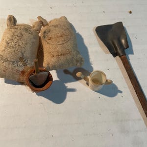 May include: A miniature gardening set featuring two small burlap sacks, a terracotta pot containing a metal hand trowel, a tiny white plastic watering can, and a long-handled metal shovel with a wooden handle, all arranged together.