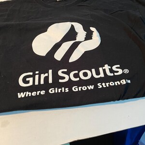 May include: A black short-sleeved t-shirt featuring a white graphic of three stylised female silhouettes. Below the graphic, the text reads Girl Scouts with the registered trademark symbol and the phrase Where Girls Grow Strong in white.