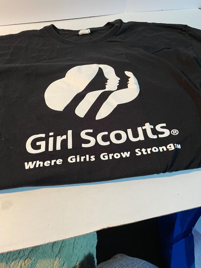May include: A black short-sleeved t-shirt featuring a white graphic of three stylised female silhouettes. Below the graphic, the text reads Girl Scouts with the registered trademark symbol and the phrase Where Girls Grow Strong in white.