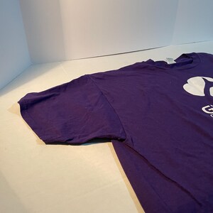 May include: A purple short-sleeved t-shirt featuring a white graphic logo on the chest. The logo includes a stylised clover shape above the partial text that reads Girl Scouts. The shirt has a crew neckline and a standard fit with visible stitching.