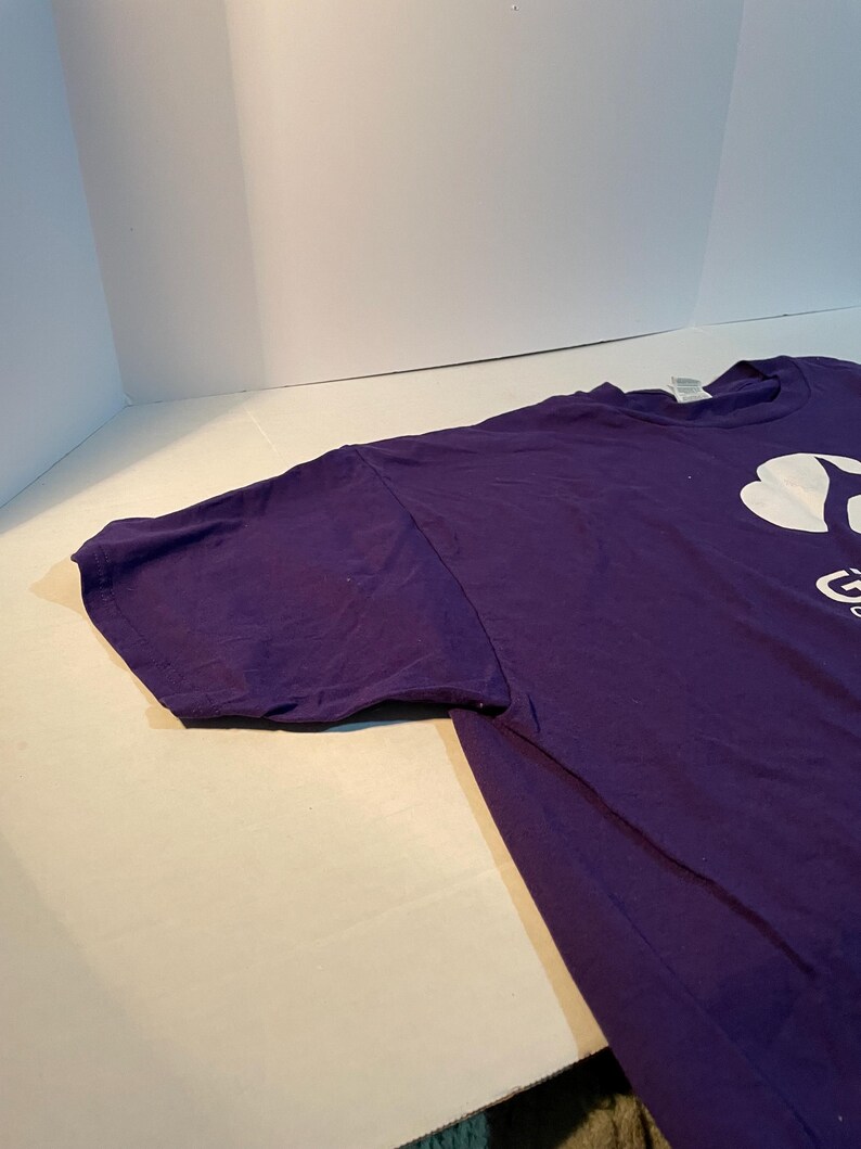 May include: A purple short-sleeved t-shirt featuring a white graphic logo on the chest. The logo includes a stylised clover shape above the partial text that reads Girl Scouts. The shirt has a crew neckline and a standard fit with visible stitching.