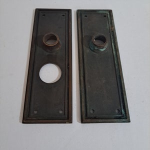 May include: Two antique brass door plates with a round hole in the centre and a smaller hole above it. The plates have a decorative border and are slightly tarnished.