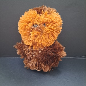 May include: A brown and orange plush toy with a furry face and a brown body. The toy has a checkered pattern on its chest.