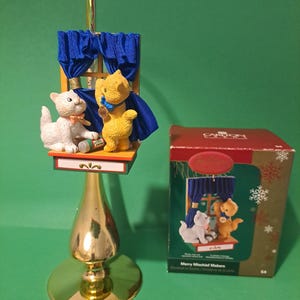Peut inclure: Ornement de Noël doré avec une scène de fenêtre représentant deux figurines de chat. Un chat blanc est assis à côté d'un chat jaune portant une cape bleue. L'ornement a une base dorée et une boîte avec "Merry Mischief Makers."