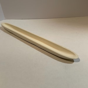 May include: A long, cream-colored ceramic dish with a shallow, rectangular design. The dish has rounded ends and a smooth, glossy finish. It is designed to hold items such as jewelry or small decorative objects.